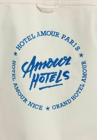 Tissu blanc avec texte brodé en bleu au centre disant "Amour Hotels", entouré par "Hotel Amour Paris", "Hotel Amour Nice" et "Grand Hotel Amour."