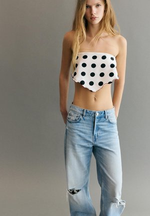 Fiatal nő fehér, pánt nélküli fekete pöttyös felsőt és világoskék, szakadt farmernadrágot visel, egy egyszerű szürke háttér előtt áll.