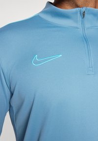 Világoskék Nike pulóver anyagból. Magas gallérral, fél cipzárral és kontrasztos, hímzett logóval a mellrészen. Sima textúra.