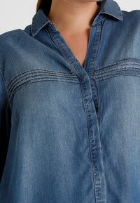 Chemise en denim avec un devant boutonné, un col classique, et des surpiqûres horizontales subtiles sur la poitrine. Couleur : bleu moyen.
