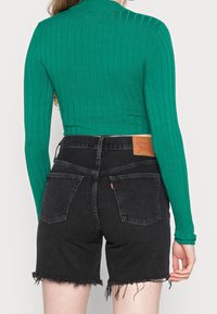 Personne portant un haut côtelé vert à manches longues et un short en jean noir taille haute avec des ourlets effilochés, vue de dos.