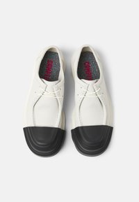 Chaussures en cuir blanc avec un capuchon en caoutchouc noir. Elles présentent des lacets fins et une semelle intérieure texturée avec des éclats multicolores. Design minimaliste.