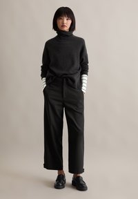 Dunkelgrauer Rollkragenpullover, lockere schwarze Hose und schwarze Schlupfschuhe. Am Bund sind weiße Streifen sichtbar. Glatte Texturen.