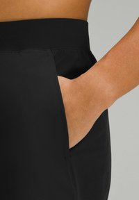 Pantalons de jogging noirs en tissu lisse et extensible, avec une poche latérale, montrant un gros plan de la main reposant à l'intérieur de la poche.
