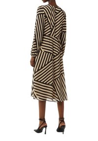 Langärmliges Kleid in Beige und Schwarz mit geometrischen Streifenmustern, tailliertem Bund und gestuftem Rock. Kombiniert mit schwarzen High Heels.