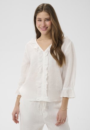 Fiatal nő hosszú barna hajjal mosolyog, fehér fodros blúzt és hozzá illő, bő szárú nadrágot visel, egyszerű világos háttér előtt.