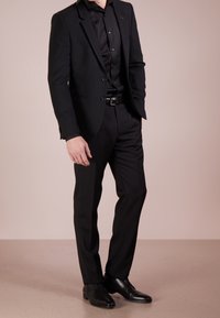 Uomo in piedi con una mano in tasca, che indossa un completo nero, una camicia nera, una cintura nera e scarpe eleganti nere su uno sfondo semplice.
