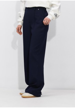 Persoon draagt wijde marineblauwe broek, wit sweatshirt met bloemenborduursel op de mouwen, en witte sneakers met gumzolen.