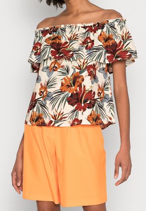Vrouw draagt een off-shoulder bloementop met rode, oranje en groene bloemen, gecombineerd met feloranje korte broek, staand tegen een effen achtergrond.