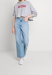 Felpa grigia cropped con logo rosso Levi's, abbinata a jeans a gamba larga blu chiaro e sneakers bianche. Realizzata in tessuto morbido e con vestibilità comoda.