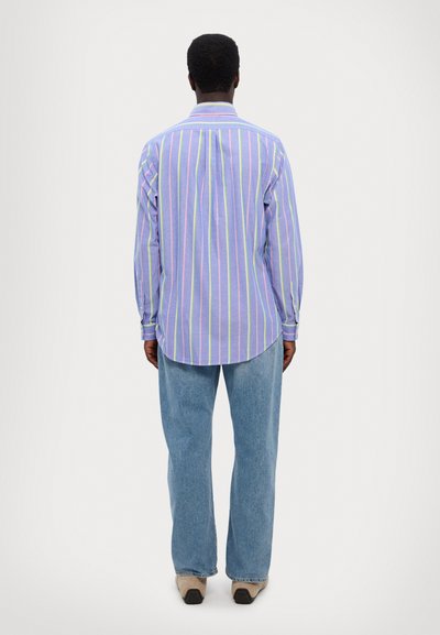 Chemise à manches longues de couleur violet clair avec des rayures verticales multicolores. Elle présente un col button-down et un ourlet arrondi. Associée à un jean bleu.