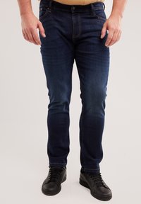 Mężczyzna ubrany w granatowe, obcisłe dżinsy oraz czarne skórzane sneakersy stoi z rękami swobodnie opartymi wzdłuż ciała.