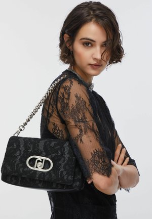 Donna con capelli corti e scuri che indossa un top in pizzo nero e porta una borsa in pizzo nero con tracolla e chiusura in catena d'argento.