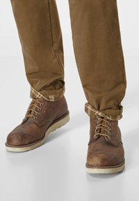 Braune Lederstiefel mit runder Zehenpartie, hellbrauner Gummisohle, goldenen Ösen und Schnürsenkeln. Die Hose hat ein kariertes Detail an der krempelbaren Fußleiste, die am Knöchel umgeschlagen ist.