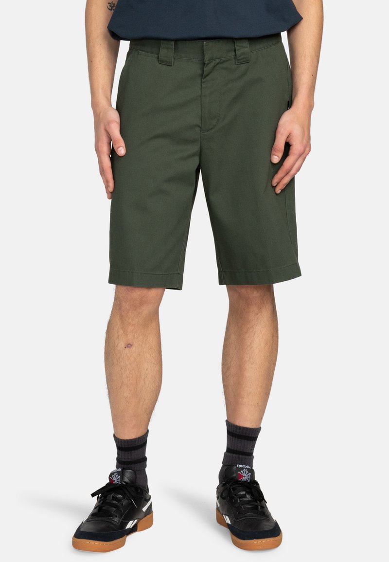 Dunkelgrüne Baumwollshorts mit flachem Bund, Seitentaschen und umsäumten Säumen. Getragen mit schwarzen Sportschuhen und dunkel gestreiften Socken.