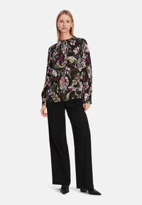 Blumenbluse mit langen Ärmeln auf schwarzem Hintergrund, mit bunten Blumen und Blättern. Dazu lockere schwarze Hose und spitze Schuhe.