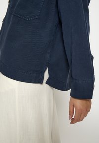 Tumši zilā krāsā pogājama krekls vieglā audumā ar sānu šķirbi un kabatu kreisajā krūtīs. Audums ar vieglām horizontālām svītrām.