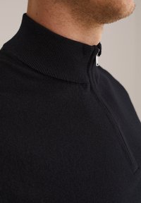 Prim-plan cu o persoană care poartă un pulover negru cu fermoar, cu guler fals texturat, închis până la gât.