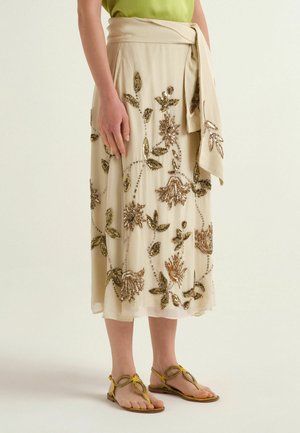 Femme portant une jupe midi beige avec une broderie florale dorée, une ceinture nouée à la taille et des sandales tressées jaunes, debout devant un fond uni.