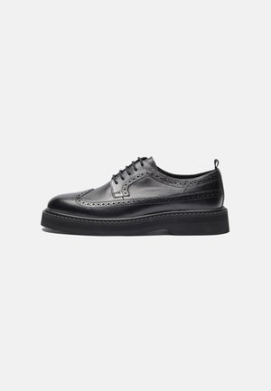 Μαύρο δερμάτινο παπούτσι τύπου brogue με σχέδιο wingtip, διάτρητες λεπτομέρειες, κλείσιμο με κορδόνια, χοντρή σόλα και έλξη στη φτέρνα, εμφανιζόμενο από το πλάι.