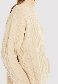 Kermanvalkoinen neulepaita, jossa on korkea kaulus, kuvioitu letinmuotoinen neulos ja rento istuvuus. Kangas näyttää pehmeältä ja paksulta kudotulta.