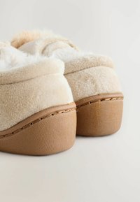 Beige, mjuka tofflor med en mjuk plyschovandel, vadderad krage och tan-färgad texturerad gummisula, med synlig sömnad längs kanten.