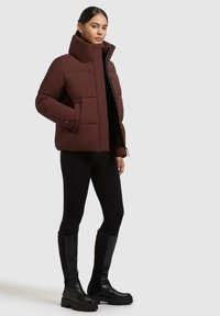Donna con giacca a vento bordeaux, leggings neri e stivali neri alti in piedi su uno sfondo grigio chiaro.