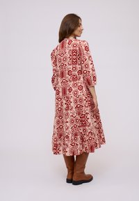 Woman in a red and cream patterned midi dress facing sideways, wearing brown knee-high boots, standing on a plain light background.