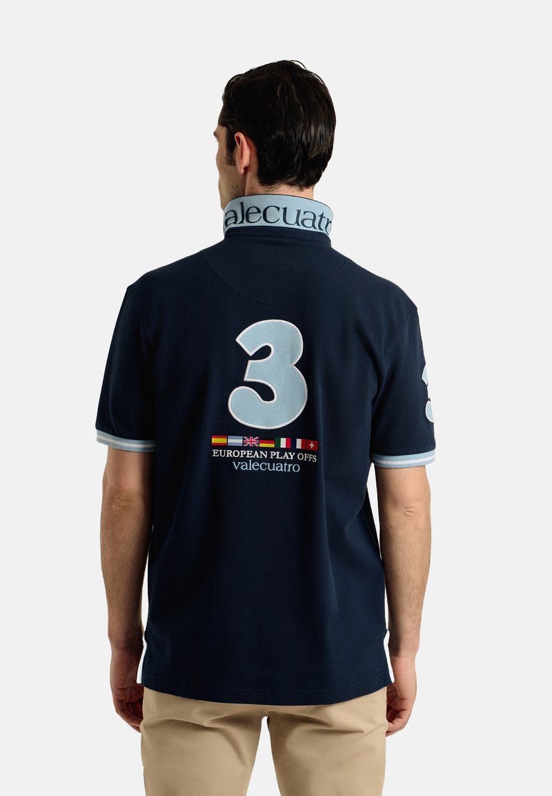 La camiseta polo azul marino presenta un gran "3" de color azul claro en la parte posterior, con texto bordado y banderas. El cuello exhibe la marca "valecuatro".