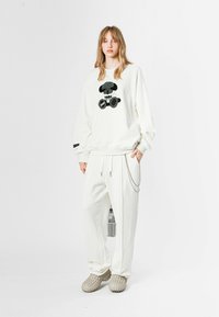 Sudadera de algodón blanca con diseño gráfico negro, combinada con pantalones a juego que tienen cordón y detalle de cadena, y zapatos beige texturizados.