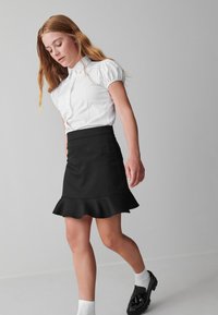 Wit shirt met korte mouwen en een kraag, gecombineerd met een zwarte rok met een uitlopende zoom. Zwarte schoenen en witte sokken maken de outfit compleet.