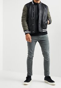 Veste bomber en cuir noir avec manches vert olive, poignets rayés et fermeture éclair à l'avant. Associée à un pantalon en denim gris et des baskets noires.
