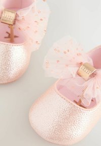 Funkelnde pinke Babyschuhe mit transparenten rosa Schleifen, die roségoldene Punkte und metallische goldene Markenanhänger auf einem neutralen Hintergrund zeigen.