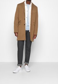 Mężczyzna w kamelowym płaszczu, założonym na białą koszulę z guzikami i jasnoniebieski sweter, ciemnych spodniach z podwiniętymi nogawkami oraz białych sneakersach na białym tle.