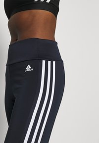 Mörkblå högmidjade leggings med tre vita vertikala ränder och en Adidas-logotyp på vänster höft. Slätt, stretchigt material.