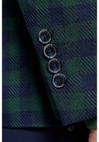 Zielona i granatowa marynarka w kratę z teksturowanym materiałem, z czterema czarnymi guzikami z białymi napisami. Zbliżenie na detale guzika.