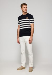 Marine gestreiftes kurzärmliges Sweatshirt mit weißen horizontalen Streifen, mit einem Brustemblem, kombiniert mit weißen Hosen und beigen Slippern.