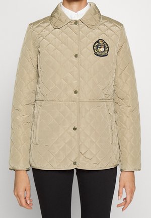 Veste matelassée beige avec boutons-pression, écusson brodé sur la poitrine, portée sur une chemise blanche, associée à un pantalon noir.
