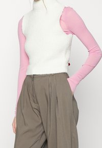 Gilet senza maniche bianco e testurizzato con colletto alto, indossato sopra una camicia a maniche lunghe rosa, abbinato a pantaloni beige a vita alta.