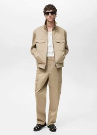 Beige Utility-Jacke mit hohem Kragen und zwei Klappentaschen, kombiniert mit passenden Cargo-Hosen und schwarzen Sandalen. Modell trägt Sonnenbrillen.