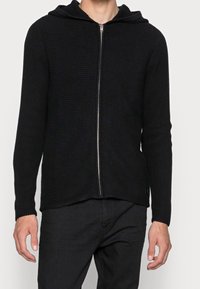 Černá mikina s kapucí na zip, vyrobená z žebrované látky. Má kapuci, dlouhé rukávy a rovný lem. Přední zip a malý detail loga na lemu.