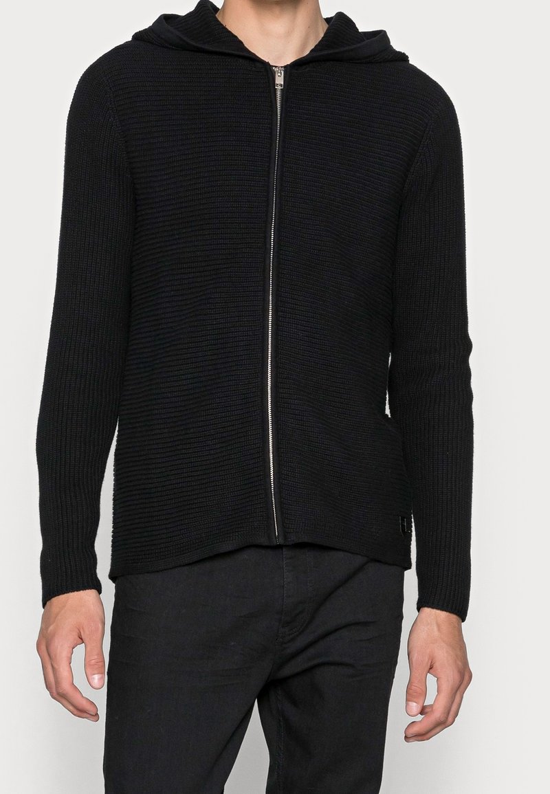 Černá mikina s kapucí na zip, vyrobená z žebrované látky. Má kapuci, dlouhé rukávy a rovný lem. Přední zip a malý detail loga na lemu.