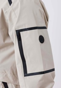 Giacca beige con dettagli neri, tasca con zip sulla manica, texture liscia e un logo circolare sul braccio.