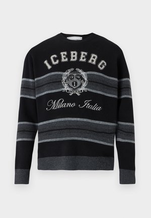 Mann trägt schwarzen und grau gestreiften "Iceberg Milano Italia" Pullover, weite dunkelgraue Hose, schwarzen Gürtel und schwarz-weiße Schuhe, steht mit einer Hand in der Tasche.