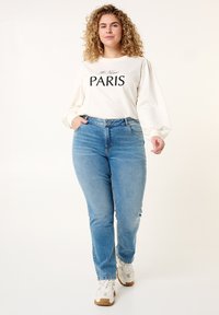 Sweat-shirt blanc avec "PARIS" imprimé en noir, assorti à un jean bleu clair et des baskets blanches avec des accents bruns. Cheveux frisés et coupe décontractée.