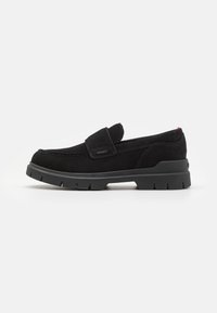 Chaussure glissante en suede noir avec un bout rond, semelle en caoutchouc noir épais et une sangle à velcro. Présente des coutures discrètes et un petit accent rouge.