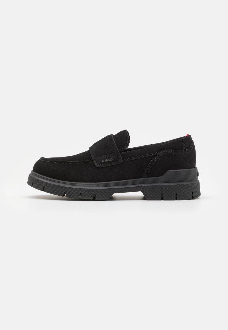 Chaussure glissante en suede noir avec un bout rond, semelle en caoutchouc noir épais et une sangle à velcro. Présente des coutures discrètes et un petit accent rouge.