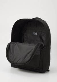 Sac à dos noir avec une fermeture éclair ouverte révélant un intérieur spacieux et une poche zippée plus petite, arborant le logo EA7.