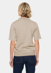 Béžový pletený polo shirt s krátkými rukávy, žebrovaným lemem a měkkou strukturou, v kombinaci s tmavými denimovými džínami, pohled zezadu.