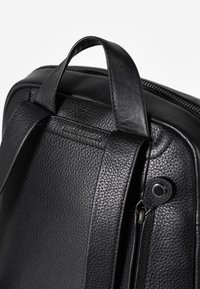 Zaino in pelle nera con superficie texturizzata, doppie spalline regolabili, manico superiore e chiusura a zip. Presenta un piccolo accento metallico.